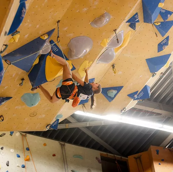 Iemand beklimt een indoor-klimwand met grote blauwe, gele en grijze grepen, terwijl hij in een uitdagende houding op een overhangend gedeelte hangt.